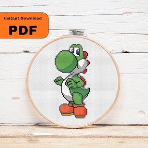May include: A cross-stitch of a green Yoshi character from the Super Mario video game series, set in a wooden embroidery hoop. The image includes the text "Instant Download PDF" in an orange rectangle.