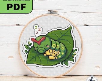 以下が含まれることがあります： 木製の刺繍枠に入ったクロスステッチパターン。赤い頭の模様がある緑色の毛虫が大きな緑の葉の上で眠っている様子が描かれています。デザインには小さな黄色い花と、緑と赤で3回繰り返された文字Zが含まれています。