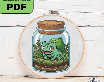 以下が含まれることがあります： 木製の刺繍枠に入ったクロスステッチパターン。背中に大きな緑の球根を乗せた明るい緑色の生き物のピクセルアートデザインで、茶色の土と緑の植物が入ったガラス瓶のテラリウムの中に座っており、コルクの蓋がされています。