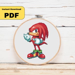 May include: A cross-stitch embroidery hoop featuring a pixelated image of Knuckles the Echidna from the Sonic the Hedgehog video game series. The character is red with white gloves and red shoes. The text "Instant Download PDF" is in a yellow rectangle.