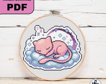 以下が含まれることがあります： 木製の刺繍枠に入ったクロスステッチの図案で、青い雲の上で眠る淡いピンク色の猫のような生き物が描かれています。デザインには紫色のアーチ、小さな青い星、そして眠りを表すために3回繰り返された文字Zが含まれています。