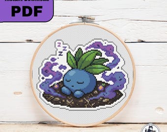 以下が含まれることがあります： 木製の刺繍枠に入ったクロスステッチパターン。頭に緑の葉を乗せて眠る青くて丸い生き物のピクセルアートデザインで、紫色の煙の雲と小さなZの文字が白いグリッドの背景に描かれています。