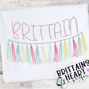 Puede incluir: Una camiseta blanca con el nombre "BRITTAIN" en letras rosa y verde, con una guirnalda de borlas de colores debajo. El texto "BRITTAINS HEART DESIGNS" está en la esquina inferior derecha.