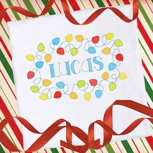 Könnte beinhalten: Weißer Strampler mit einem bunten Lichterketten-Design und dem Namen "Lucas", der in Blau gestickt ist.