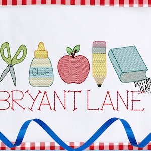 May include: Embroidered design featuring a red apple, scissors, glue bottle, pencil, and a blue book with the name "BRYANT LANE" in red thread. The design is on a white background with a red and white checkered border.