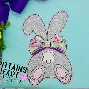 May include: A gray bunny with a floral bow, embroidered on a light blue fabric. The bunny is facing away from the viewer, with its tail and two feet visible. The text "Brittains Heart Designs" is embroidered in black on the fabric.