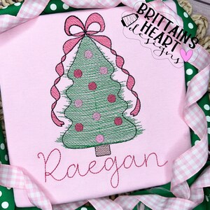 May include: Pink t-shirt with an embroidered Christmas tree design. The tree is green with pink ornaments and a pink bow at the top. The name "Raegan" is embroidered in pink script below the tree. The shirt is framed with pink and white ribbon.