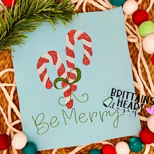 Könnte beinhalten: Stickerei mit zwei roten und weißen Zuckerstangen, die mit einem grünen Band zusammengebunden sind. Der Text "Be Merry" ist in Grün unter den Zuckerstangen gestickt.