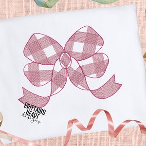 Puede incluir: Tela blanca con un bordado de lazo a cuadros rosa y blanco. El lazo tiene un patrón de puntadas detallado. El texto "BRITTAINS HEART designs" está impreso en negro debajo. Cintas en tonos dorados y rosas en el fondo.