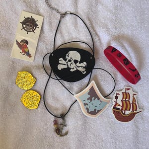 May include: A collection of pirate-themed accessories, including a black eye patch with a skull and crossbones, a red bracelet, gold coins, and a necklace with an anchor charm. Also included are stickers of a parrot, a map, and a ship.