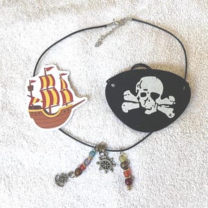May include: A pirate-themed necklace featuring a black cord with a ship-shaped charm, a black eye patch with a skull and crossbones, and beaded accents. The necklace includes a silver-colored ship wheel charm and colorful beads.