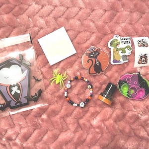 May include: A collection of Halloween-themed items including a clear plastic bag with a vampire graphic, a small neon green plastic spider, a beaded bracelet with a skull charm, a black cat sticker, a zombie sticker labeled SPOOKY TIME, and a pumpkin toy.