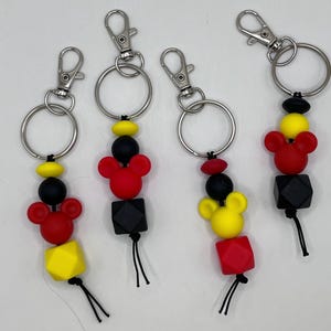 May include: Four keychains with silver rings and lobster claw clasps. Each keychain features a beaded design with red, yellow, and black beads, including Mickey Mouse-shaped beads and geometric shapes. Black tassels hang from the bottom.
