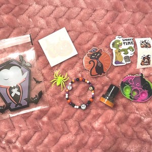 May include: A collection of Halloween-themed items, including stickers, a bracelet, a pop-it toy, and a bag with a vampire coffin design. The items are in various colors, such as orange, black, purple, and green, and are arranged on a pink surface.