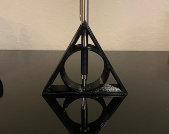 May include: A black 3D printed pen holder shaped like the Deathly Hallows symbol from the Harry Potter series. A silver pen is standing upright in the holder.