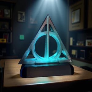 May include: A light-up, silver-colored Deathly Hallows symbol, a triangle with a circle and a vertical line, sits on a dark brown rectangular base. The symbol glows with a bright blue light. The item is on a wooden table.