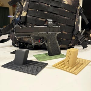 Może przedstawiać: Czarny pistolet z czerwonym celownikiem punktowym, spoczywający na czarno-zielonym stojaku 3D w kształcie amerykańskiej flagi. Na zdjęciu znajduje się również beżowy stojak 3D w kształcie amerykańskiej flagi z literami "USA". W tle znajduje się plecak w kamuflażu.