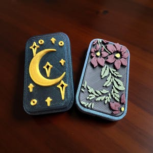 May include: Two rectangular cases with raised designs. One features a gold crescent moon and stars on a dark blue background. The other has purple flowers and green leaves on a dark background. Both have a textured appearance.