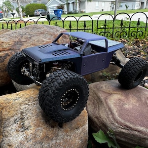 May include: A blue and black remote control truck with a black roll cage and large black tires. The truck is sitting on a large rock.