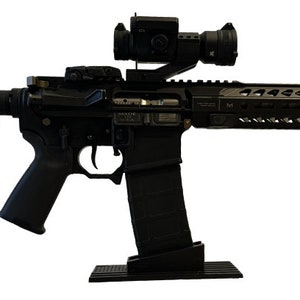 Puede incluir: Un rifle negro con múltiples accesorios, incluyendo una mira telescópica, una linterna y un cargador. El rifle tiene una culata y empuñadura negras. La imagen está sobre un fondo blanco.