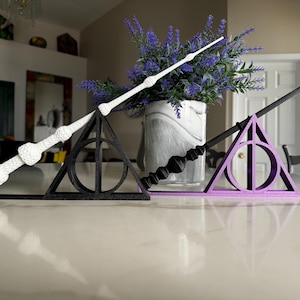 May include: Two black and white wands on a black and purple triangle stand. The wands are on a white surface. The stand is shaped like the Deathly Hallows symbol.