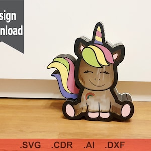 Puede incluir: Un recorte de madera de un unicornio con una melena y cola de arcoíris. El unicornio está sentado con los ojos cerrados y tiene un arcoíris en un lado. El unicornio está pintado en tonos de marrón, rosa, amarillo y verde.