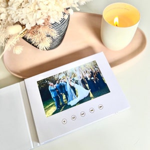 Op de afbeelding: Een wit fotoalbum met een ingebouwd scherm waarop een trouwfoto te zien is van een bruid en bruidegom met hun bruiloftsgezelschap. Het album heeft gouden knoppen om af te spelen, te pauzeren en terug te spoelen.
