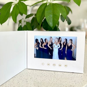Op de afbeelding: Een wit fotoalbum met een ingebouwd scherm waarop een groep vrouwen in zwarte en witte jurken te zien is. Het scherm heeft afspeelknoppen.