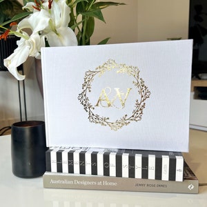 Op de afbeelding: Een wit hardback boek met een gouden folieontwerp van een krans met de initialen "A&W" in het midden. Het boek ligt opgestapeld op twee andere boeken, een met zwart-witte strepen en de tekst "Elements of Style" en de andere met de tekst "Australian Designers at Home" en "Jenny Rose-Innes".