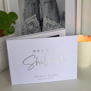 Op de afbeelding: Wit bruiloftsgastenboek met zilveren folie opschrift "MR + MRS Shilvock" en "THE MILL BARNS 02.07.2023".