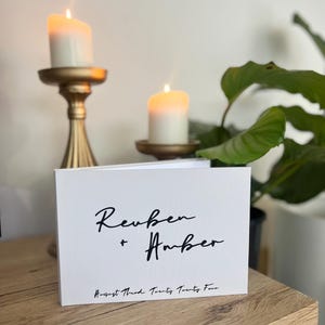 Op de afbeelding: Witte kaart met zwarte kalligrafie die "Reuben + Amber" en "August Third, Twenty Twenty Four" luidt. De kaart ligt op een houten oppervlak met twee brandende kaarsen en een groene plant op de achtergrond.
