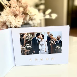 Op de afbeelding: Een wit fotoalbum met een scherm waarop een trouwfoto te zien is. De afbeelding toont een bruid en bruidegom die kussen, omringd door bruiloftgasten. Gouden afspeelknoppen zijn zichtbaar aan de onderkant van het scherm. Droogbloemen staan op de achtergrond.