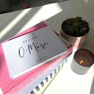 Op de afbeelding: Een witte kaart met zwarte tekst die "MR & MRS O'Mara 28.08.23" zegt, op een stapel boeken. Het bovenste boek is roze, het volgende is blauw met witte strepen en het onderste boek is zwart met witte strepen. Een kleine pot met een vetplant en een kaars staan op de tafel.
