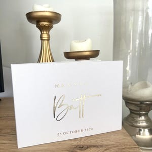 Op de afbeelding: Wit bruiloftsgastenboek met gouden folielettering. De tekst luidt "Mr. & Mrs. Batt" en "05 oktober 2024".