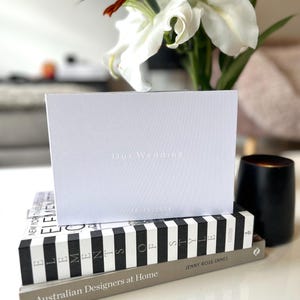Op de afbeelding: Een witte kaart met de tekst "Our Wedding" en de datum "28.10.2024" erop gedrukt. De kaart ligt op een stapel boeken.