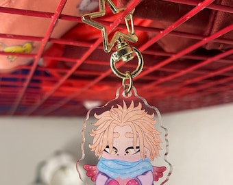 May include: A clear acrylic keychain featuring a cartoon character with spiky blonde hair, a light blue scarf, and small red wings. The character is wearing a purple outfit and holding a pink object. It has a gold-colored star-shaped metal clasp.