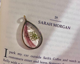 May include: A close-up of a small, clear, teardrop-shaped pendant with a delicate, dried flower pressed inside. The pendant is made of metal and has a gold finish. The flower is white with a hint of pink and is surrounded by a thin, red stem.