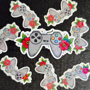 May include: A collection of gray video game controller stickers, each adorned with vibrant red flowers and green leaves. The controllers feature colorful buttons and directional pads, creating a playful and artistic design. The stickers are scattered on a dark surface.