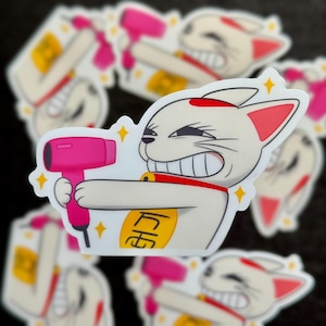May include: A white cat with a red spot on its head is holding a pink hairdryer. The cat has a gold coin around its neck with the Japanese character for "fortune" written on it. The cat is smiling with its teeth showing. The background is a black surface with other stickers of the same cat.