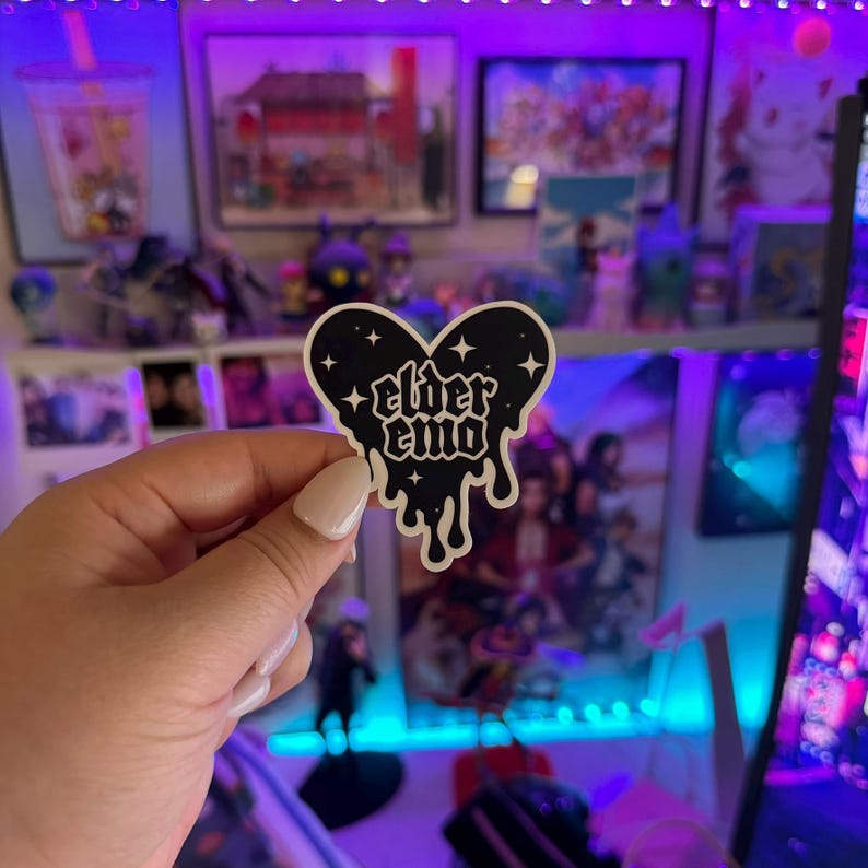 May include: A black and white sticker in the shape of a dripping heart with the text "elder emo" inside. The sticker has a starry background.