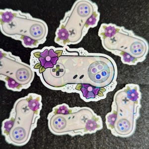 May include: A set of eight stickers featuring a retro video game controller with purple flowers. The stickers are holographic and have a white background.
