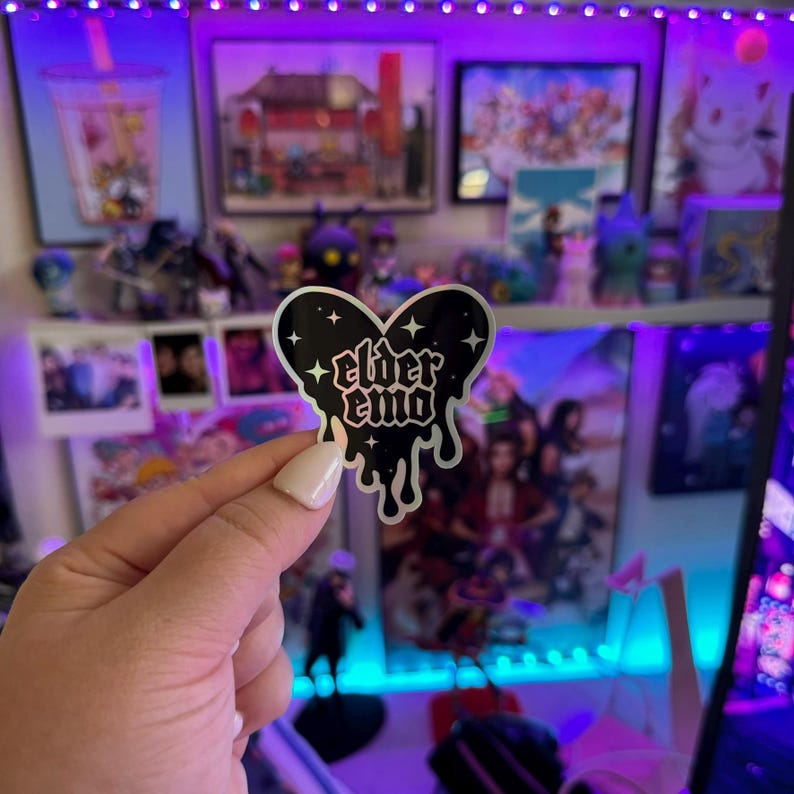 May include: A black and silver heart-shaped sticker with the text "elder emo" in a gothic font. The sticker has a dripping effect and stars around the edges.
