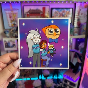 May include: Square sticker featuring cartoon characters from The Owl House. The characters include Eda, Luz, King, and Hooty. They are wearing blue sweaters against a purple background with diamond shapes. The sticker is held by a person.