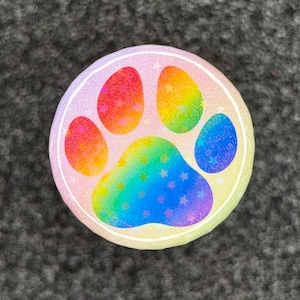 May include: A round, rainbow-colored paw print design with a white outline. The paw print features a gradient of colors, including red, orange, yellow, green, and blue, with small star accents. The background is a light pink.