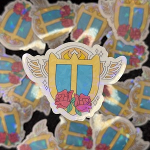 May include: A holographic sticker featuring a gold shield with a blue center, two wings, and two pink roses. The shield has a gold crown on top and a gold border.