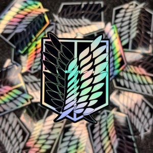 May include: A holographic sticker featuring the iconic Wings of Freedom symbol from the anime Attack on Titan. The symbol is a black and white silhouette of two wings with a rectangular frame around them. The sticker has a rainbow iridescent finish.