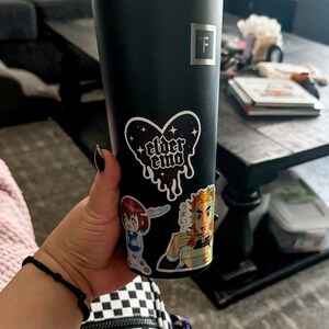 May include: A black stainless steel tumbler with a clear plastic straw. The tumbler has a sticker of a heart with the words "elder emo" on it. There are other stickers on the tumbler, including one of a cartoon character.