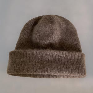 Könnte beinhalten: Eine braune Strickmütze mit umgeschlagenem Rand. Die Mütze besteht aus einem weichen, strukturierten Material und hat eine einfarbige, neutrale Farbe. Diese Wintermütze eignet sich für kaltes Wetter und Freizeitkleidung.