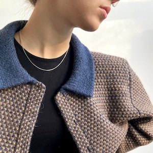 Puede incluir: Una chaqueta corta de color marrón y azul con un patrón de cuadros y un cuello azul. La chaqueta tiene cierre de botón y está hecha de una tela suave y texturizada.