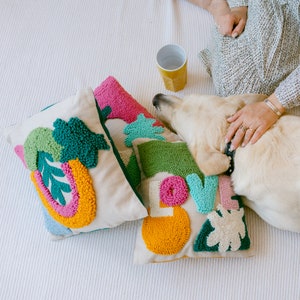 Könnte beinhalten: Drei bunte Kissen mit verschiedenen Texturen und Mustern. Die Kissen sind in einem Mix aus Grün, Pink, Gelb und Blau gehalten. Die Kissen liegen auf einer weißen Oberfläche, auf der ein Hund schläft.